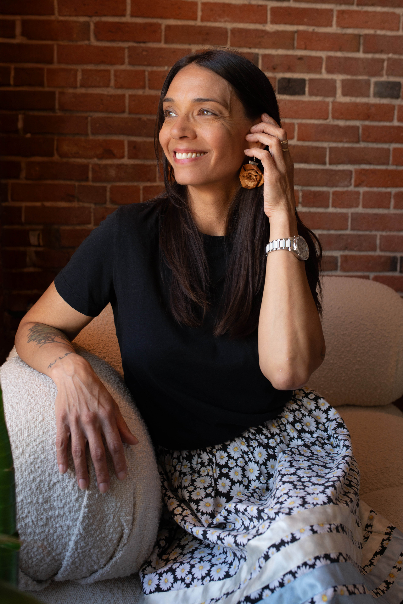 Midlife woman with long dark hair and a wooden rose earring, bright smile against a warm brick wall